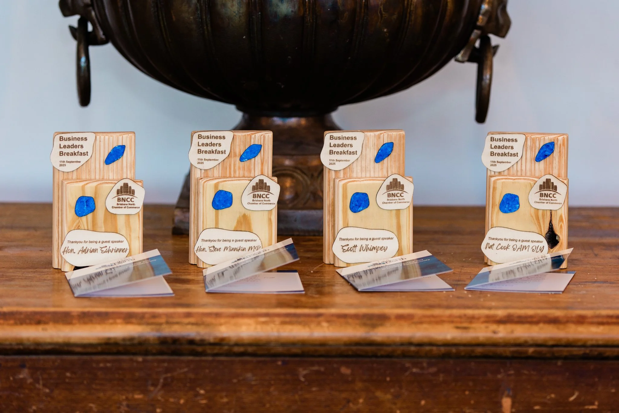 Four 'Business Leaders Breakfast' trophies engraved for guest speakers including the Hon. Adrian Schrinner, displayed on a timber sideboard.