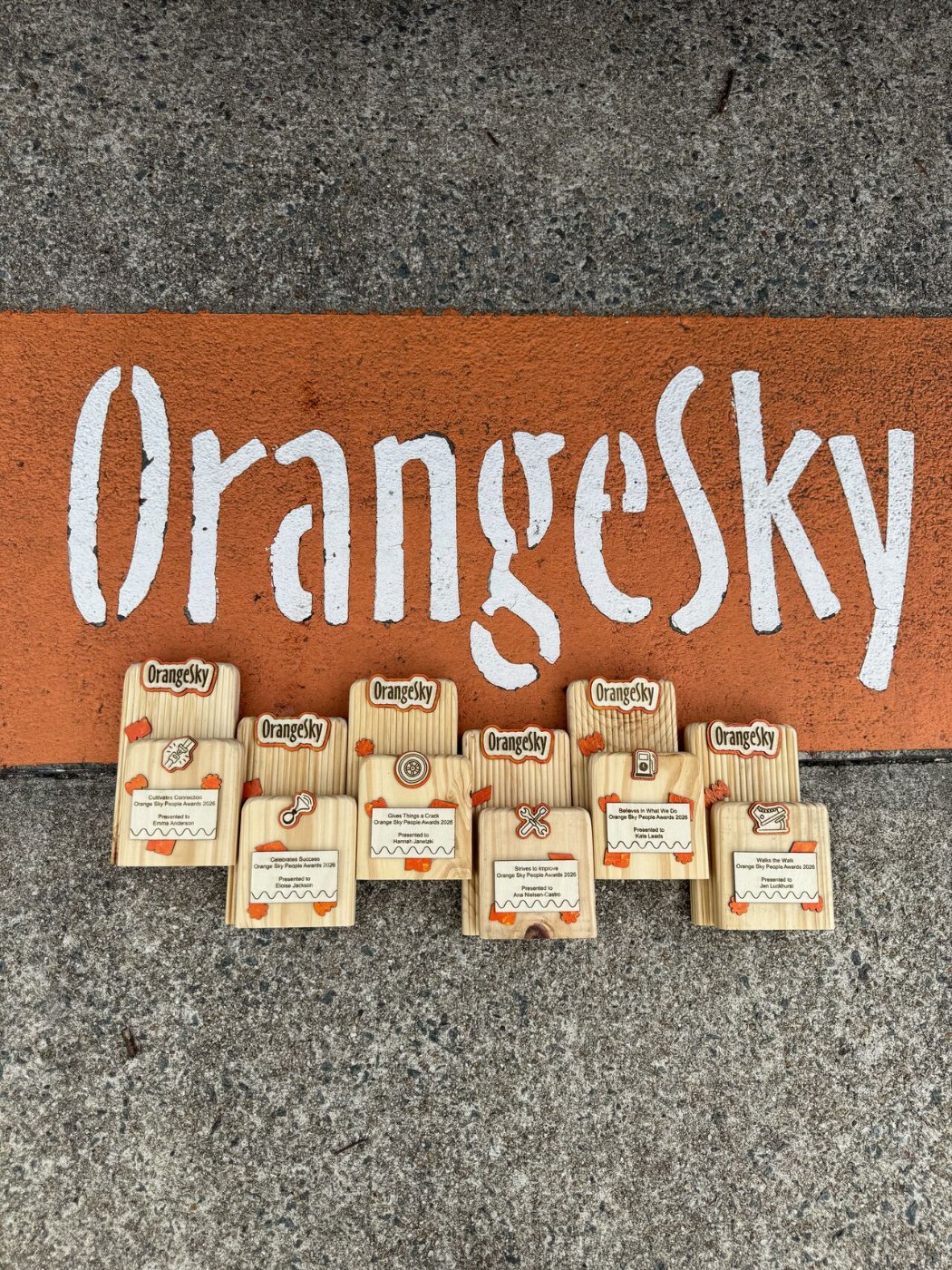 A full set of Orange Sky 'People Awards 2026' trophies in pine with the orange Orange Sky logo, arranged beneath a painted Orange Sky mural.