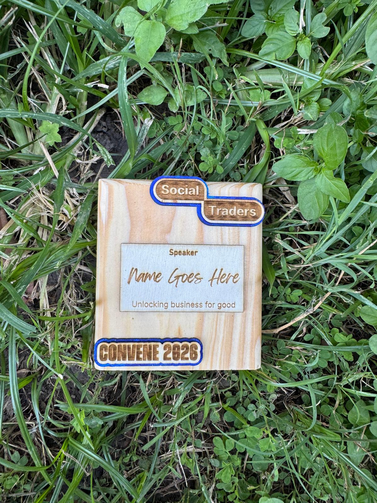 A reclaimed-timber conference speaker piece engraved 'Speaker — Name Goes Here, Unlocking business for good' with a Social Traders and CONVENE 2026 lockup.