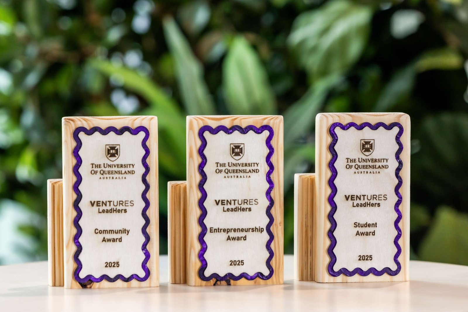 Three University of Queensland 'VENTURES LeadHers' awards in reclaimed timber with hand-poured purple wavy borders, set against tropical greenery.
